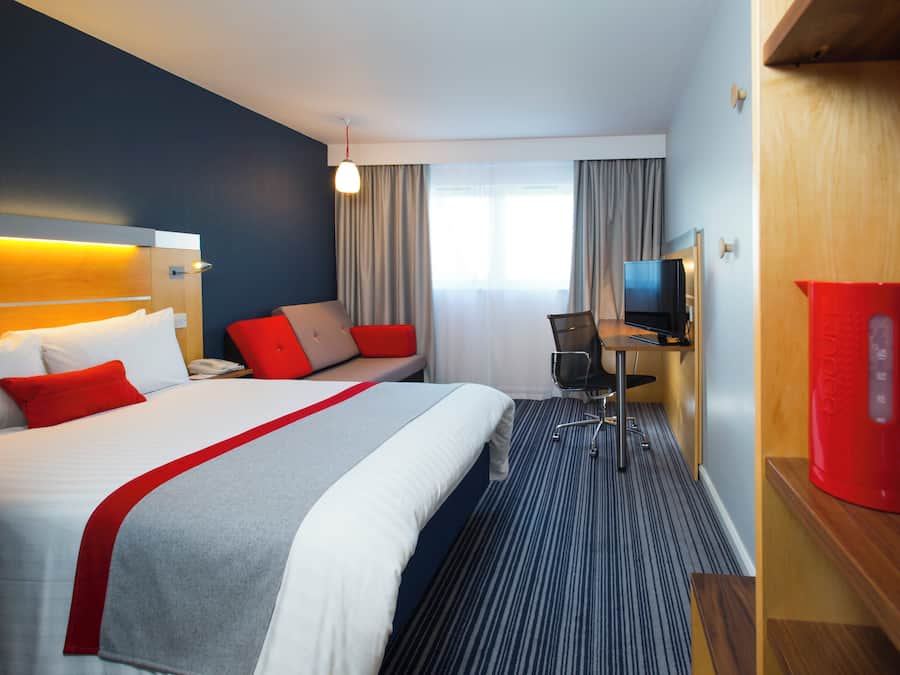 Et hotelværelse med seng, skrivebord, stol, fjernsyn og en rød højttaler.