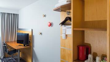 Habitación de hotel con escritorio, televisión, armario, tetera roja y estante con artículos de aseo.