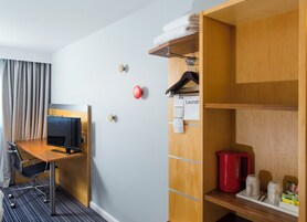 Habitación de hotel con escritorio, televisión, armario, tetera roja y estante con artículos de aseo.
