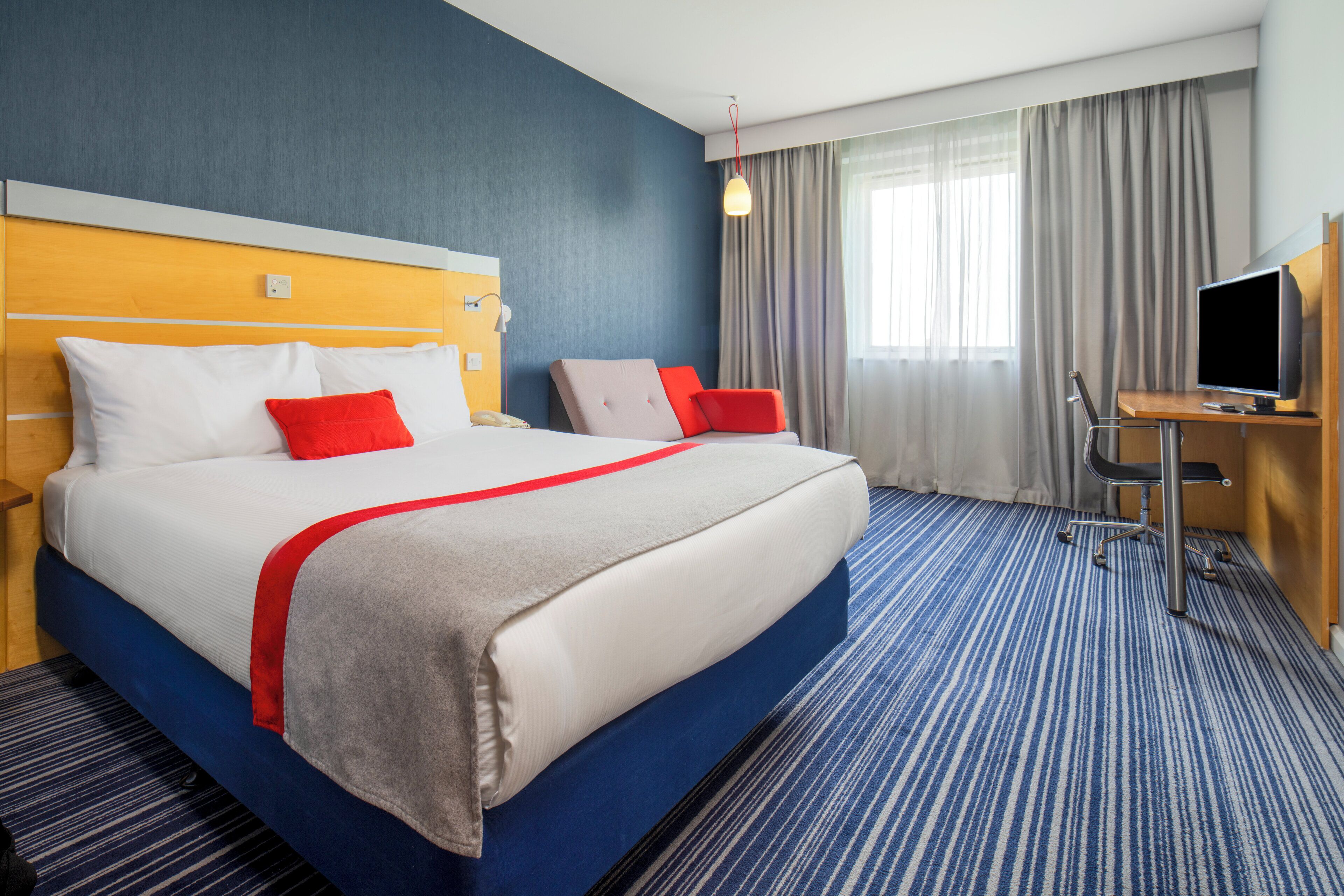 A hotel room with a large bed, a desk with a TV, and a window with curtains.