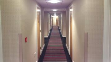 A hotel hallway with carpeted floors, doors on both sides, and a fire alarm on the left wall.