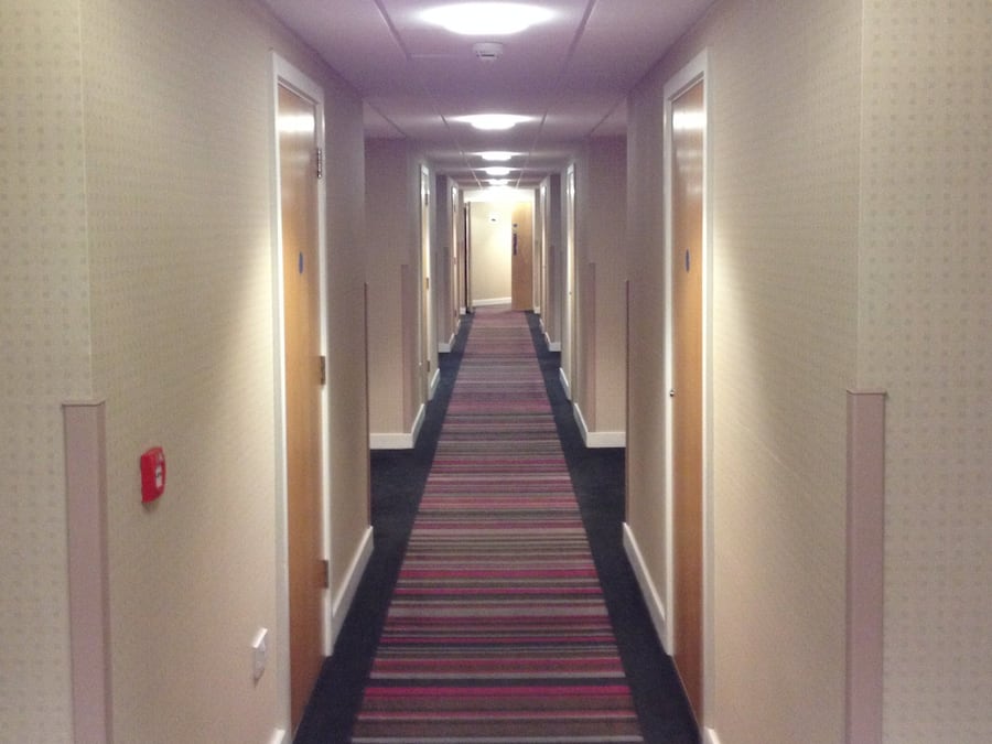 A hotel hallway with carpeted floors, doors on both sides, and a fire alarm on the left wall.