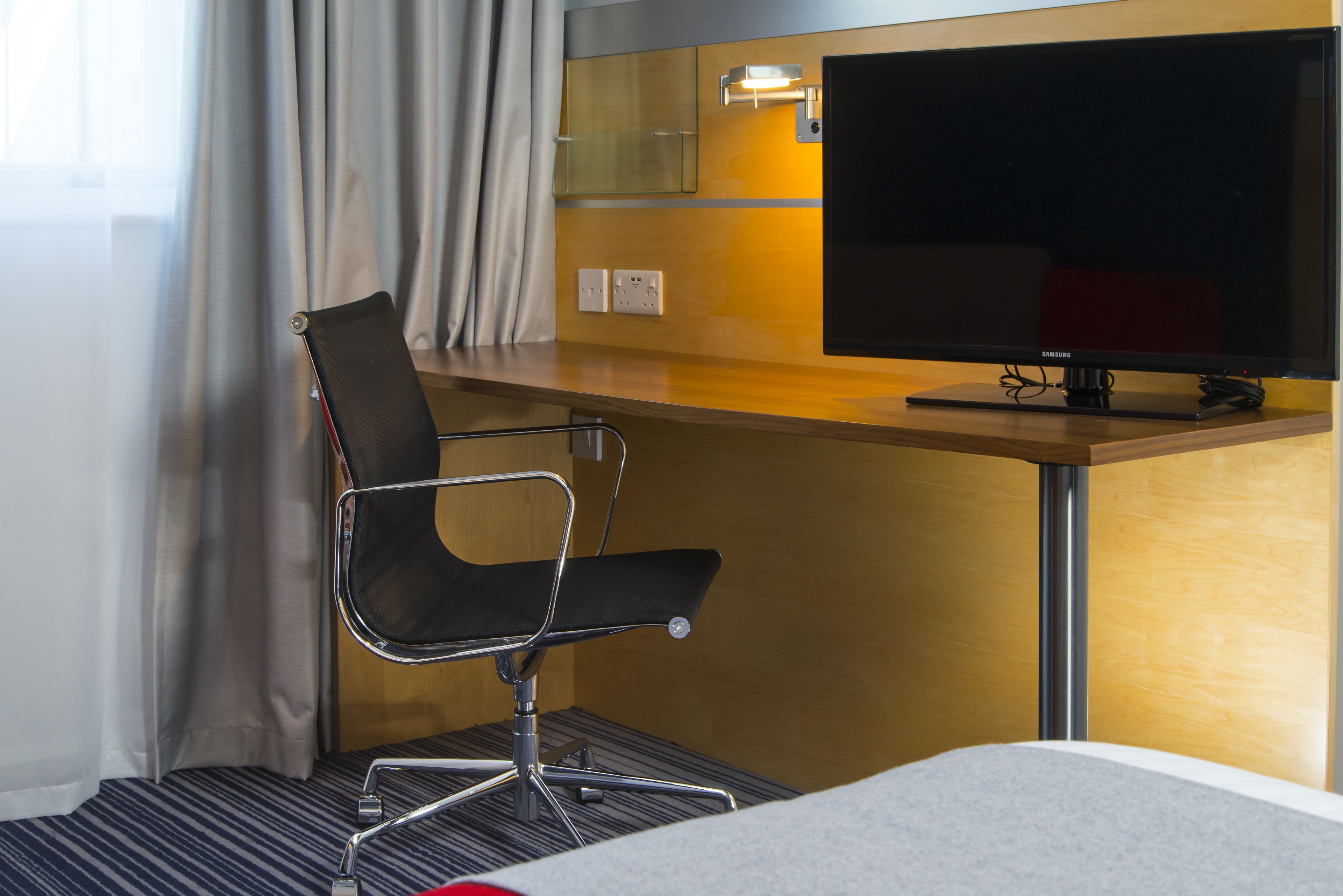 A hotel room with a bed, a desk with a television, a chair, and a window with curtains.