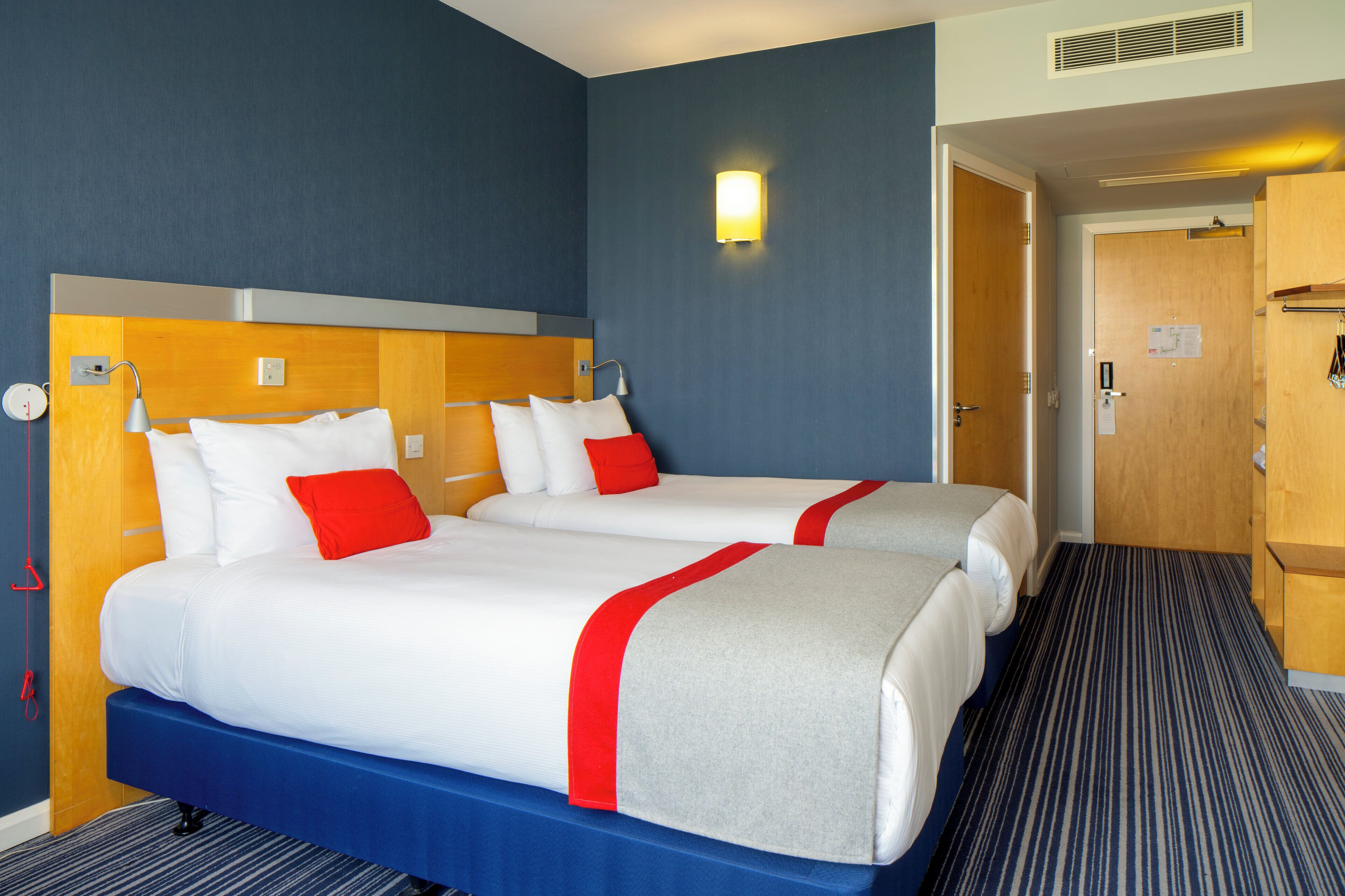 Habitación de hotel con dos camas, cabecero de madera, colcha roja y blanca, alfombra azul, y lámpara de pared.