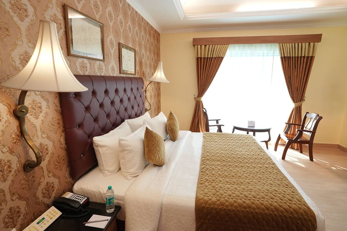 Habitación de hotel con cabecera capitonada, una cama grande con ropa de cama blanca y marrón, dos lámparas de noche, una mesita de noche con un teléfono y una botella de agua, una pequeña mesa con una silla y vistas al exterior a través de cortinas.