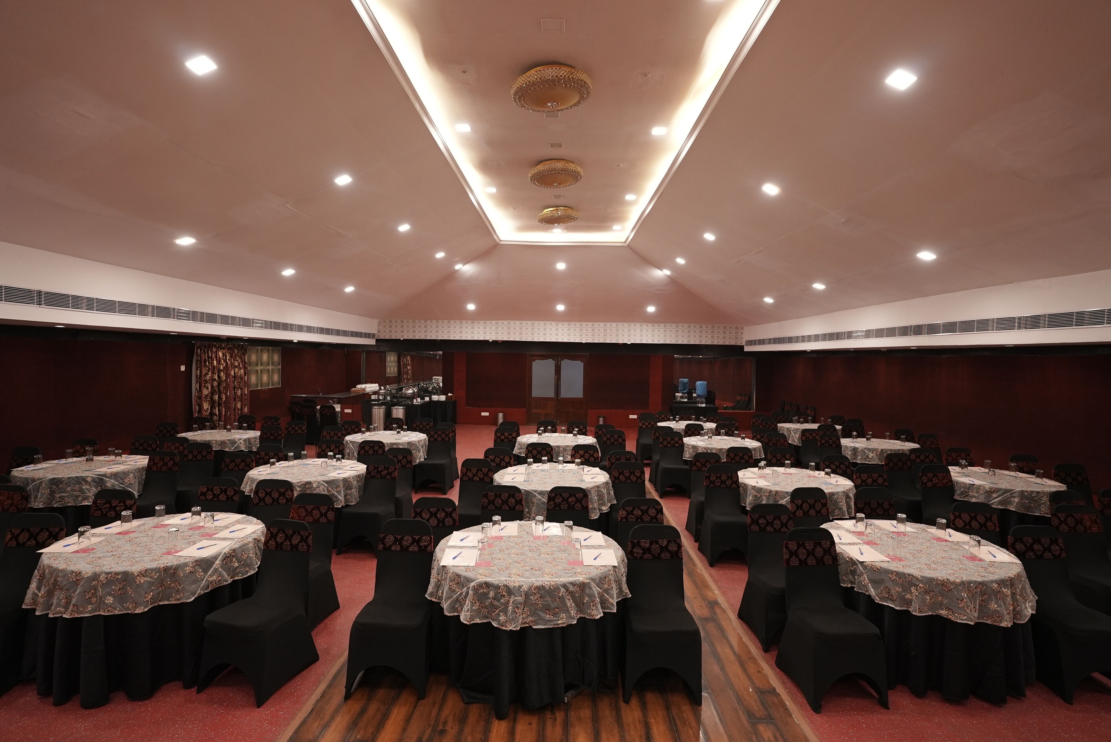 A banquet hall with round tables covered with lace tablecloths, black chairs, and a high ceiling with recessed lighting.