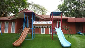 Un parque infantil con dos toboganes y una estructura para escalar, frente a un edificio de techo rojo y ventanas.