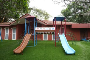 Un parque infantil con dos toboganes y una estructura para escalar, frente a un edificio de techo rojo y ventanas.