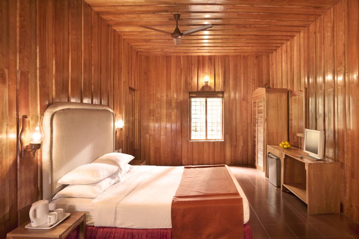 Een kamer met houten lambrisering, een bed, een bureau met een televisie en een raam met luxaflex.