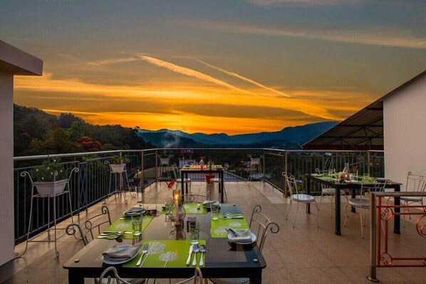 Een terras op het dak met eettafels, met uitzicht op een bergachtig landschap tijdens de zonsondergang.