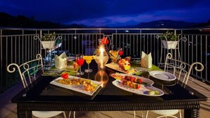 Une table à manger dressée avec différents plats et boissons sur un balcon offrant une vue sur les montagnes au crépuscule.