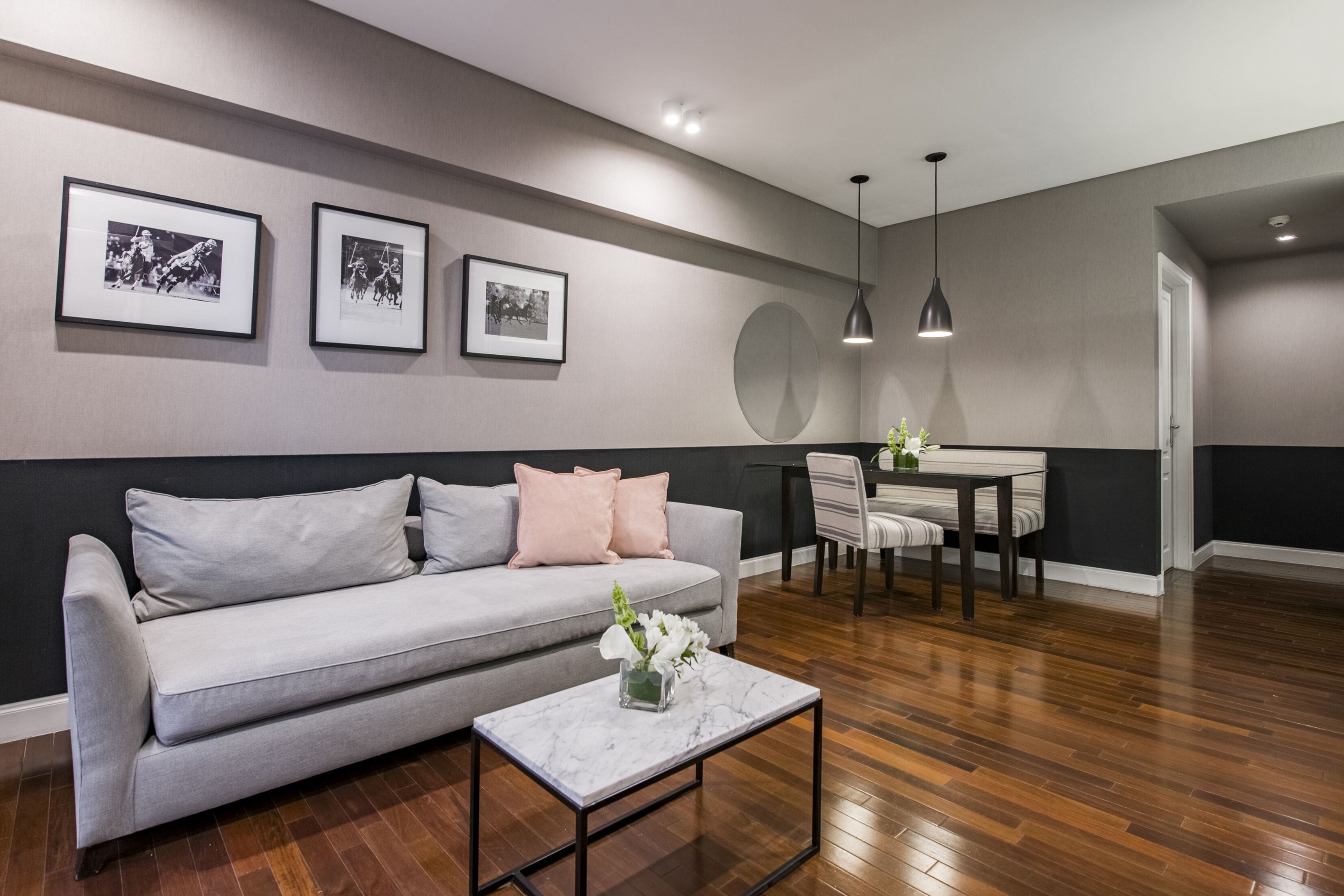 Un salon moderne avec un canapé gris, une petite table basse ornée d’un vase de fleurs, un coin repas avec une table et des chaises, et des tableaux encadrés accrochés au mur.