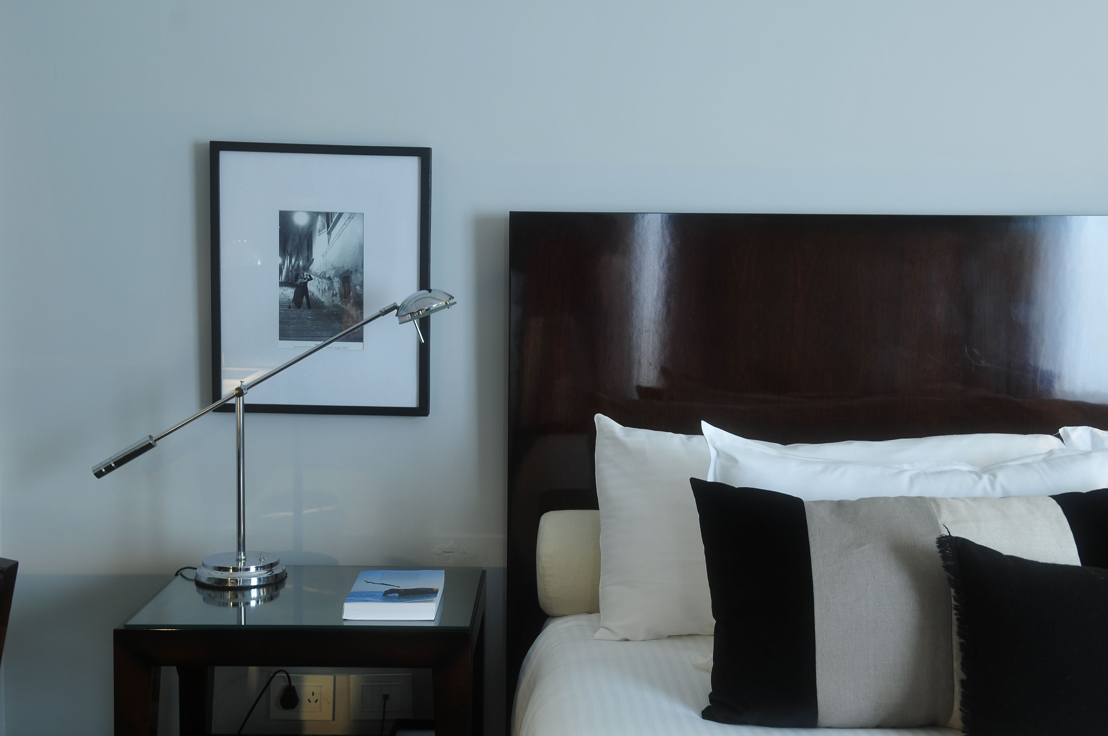 Una cama bien tendida con almohadas blancas y negras, un cuadro en la pared y un escritorio con una lámpara y un libro.