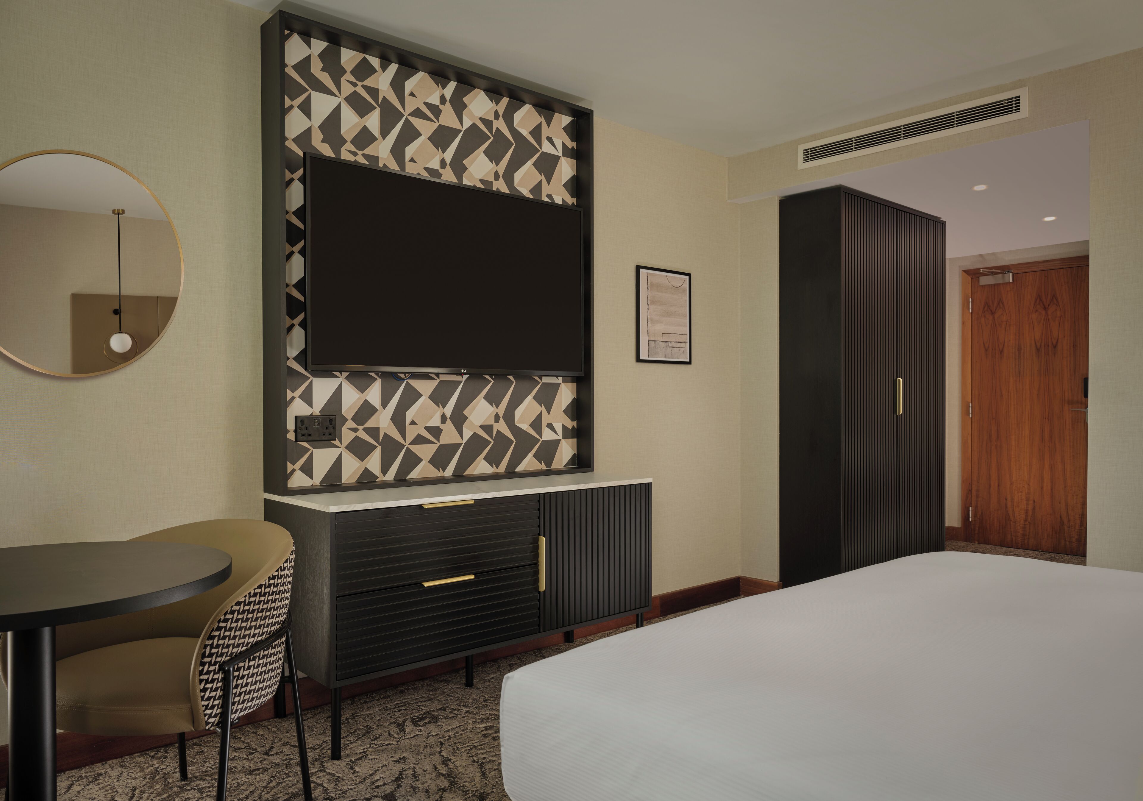 A hotel room with a bed, a desk with a chair, a TV mounted on the wall, a wardrobe, and a door.