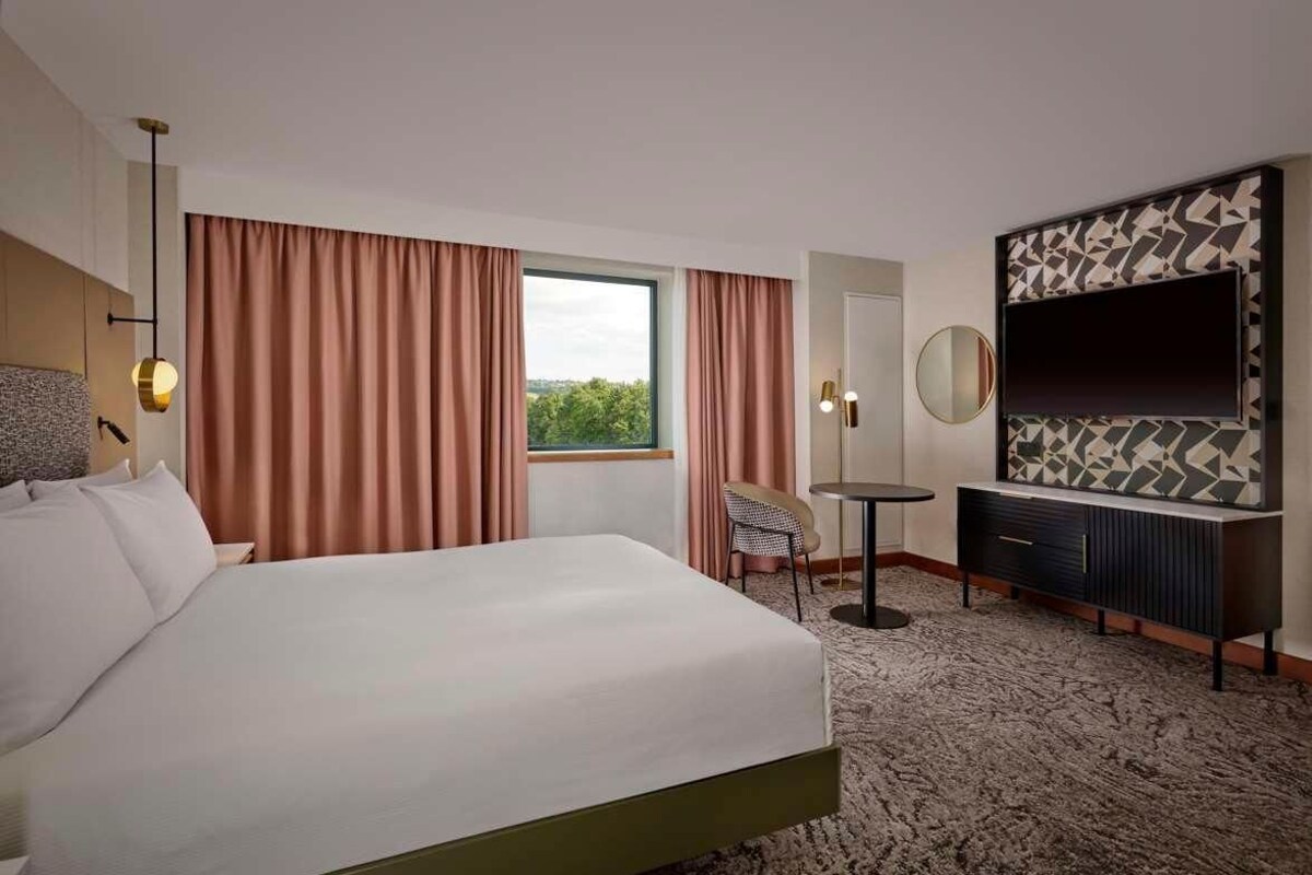 A hotel room with a bed, a TV, a small table, a chair, and a window with curtains.