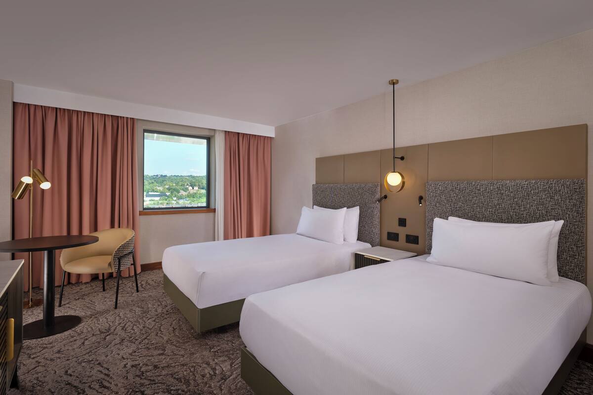 A hotel room with two beds, a desk with a chair, a large window with curtains, and a view of the city.