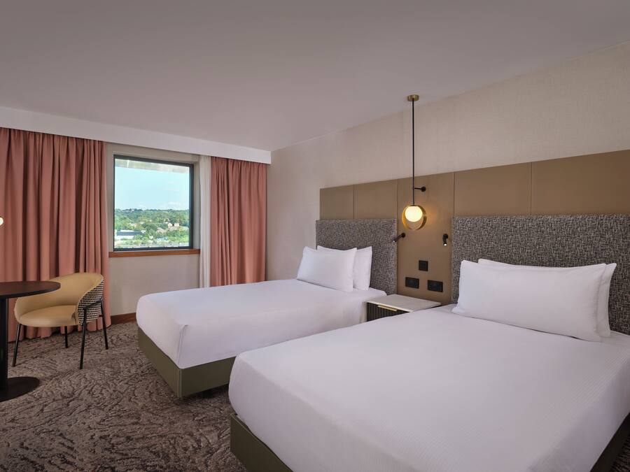 A hotel room with two beds, a desk with a chair, a large window with curtains, and a view of the city.