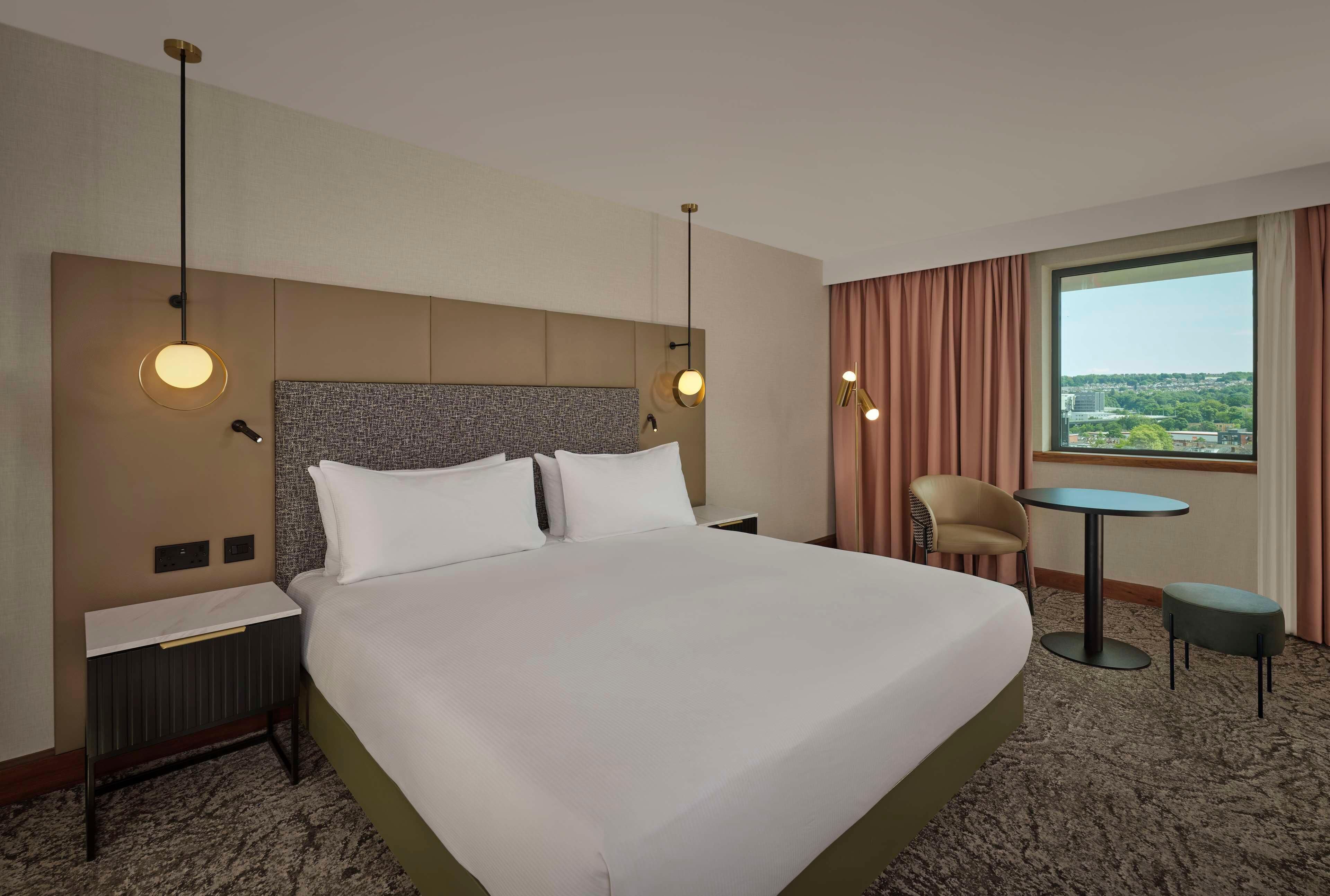 A hotel room with a large bed, a nightstand, a chair, a small table, and a view of the city.