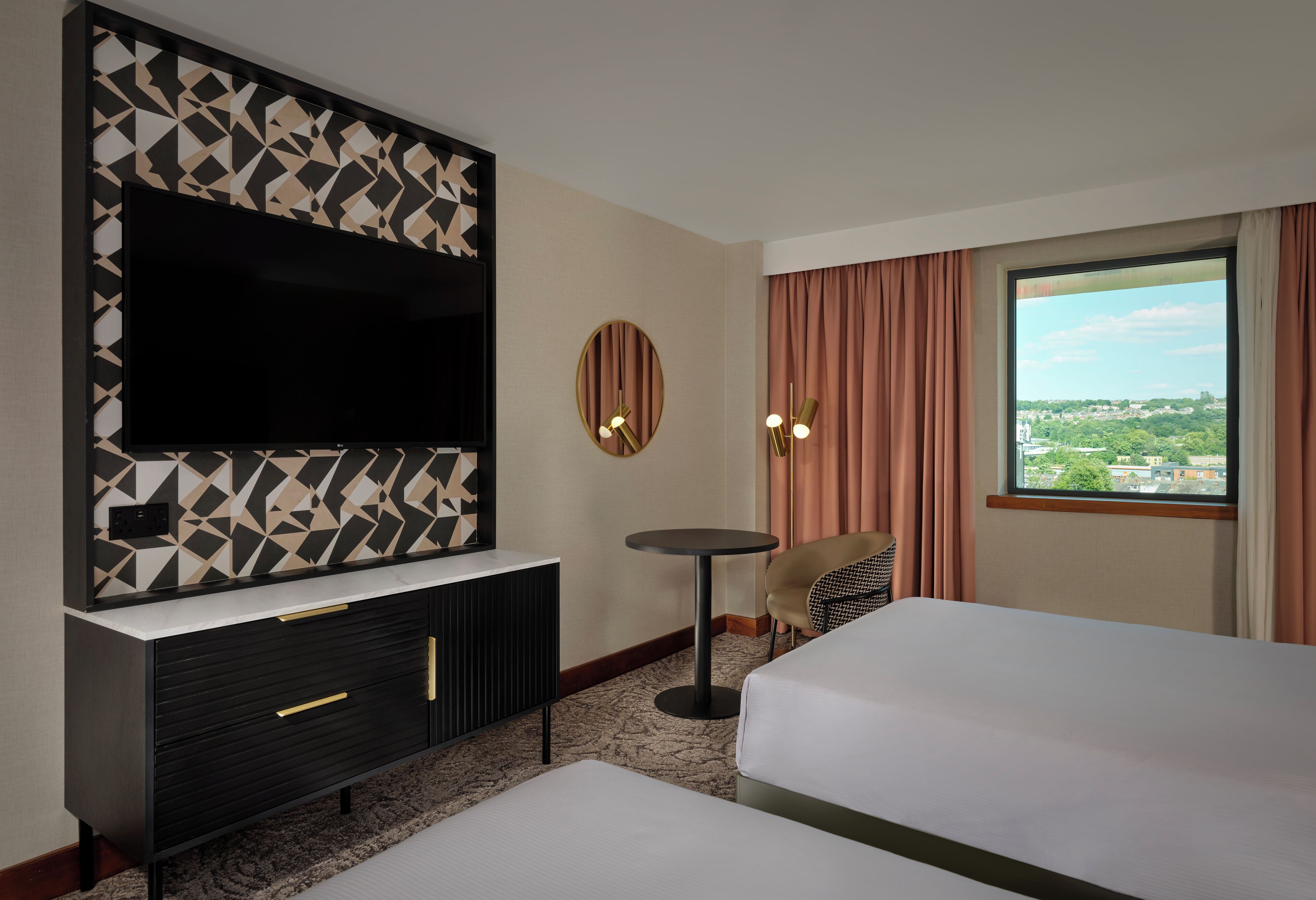 A hotel room with a large flat-screen TV, a round mirror, a small round table, a bed, and a view of the city through a window.