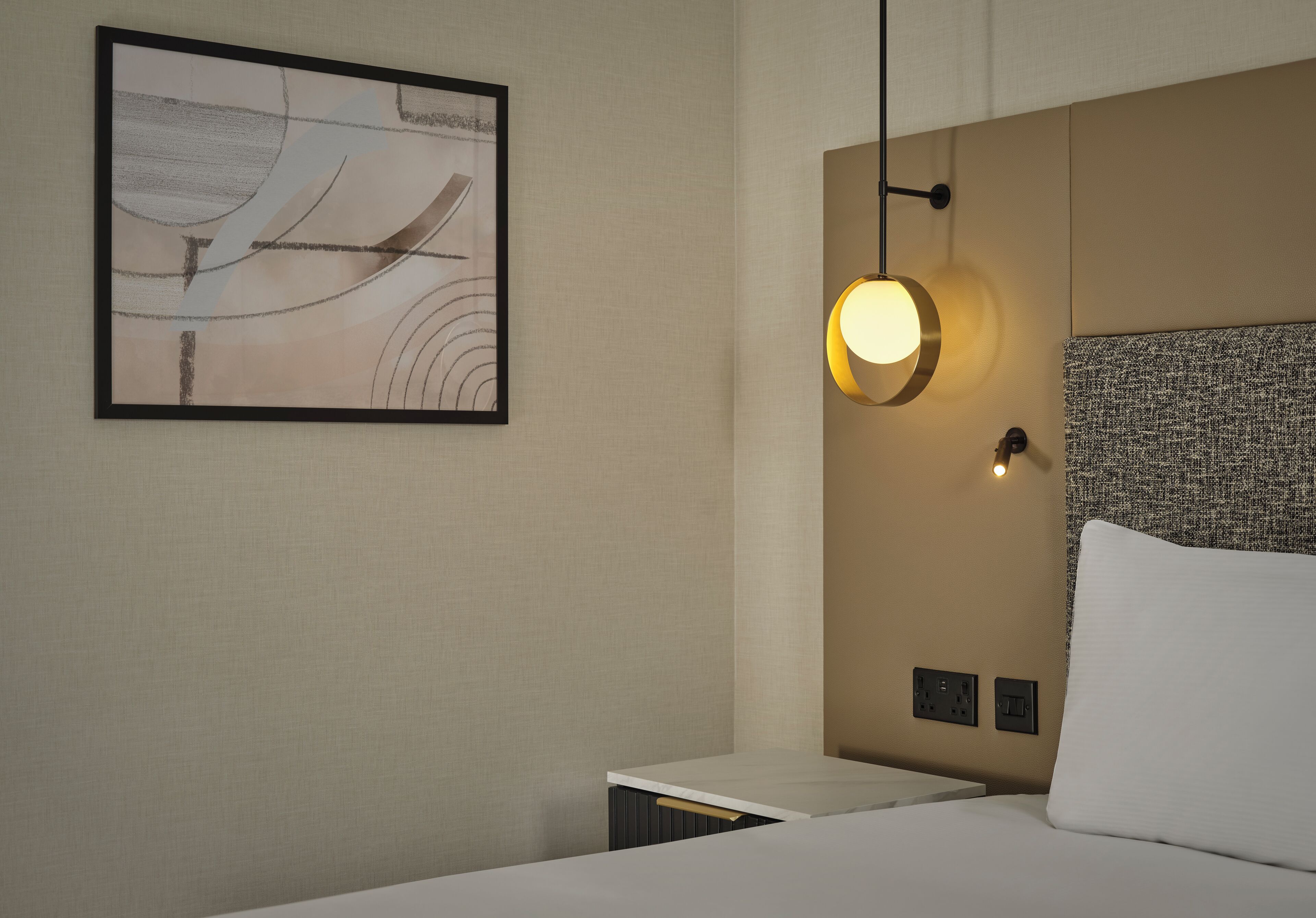 A hotel room with a bed, a bedside table, a wall-mounted light, and a framed artwork.