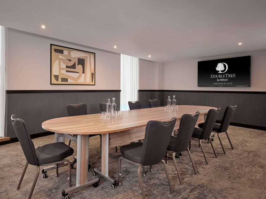 Ein Konferenzraum mit einem großen ovalen Tisch, sechs Stühlen und einem Fernsehbildschirm, der das Logo von DoubleTree by Hilton zeigt.