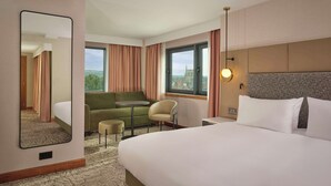 A hotel room with a large bed, a sofa, a small table, a chair, and a large mirror. There is a window with curtains and a view of a building outside. - DoubleTree by Hilton Sheffield City (Sheffield)
