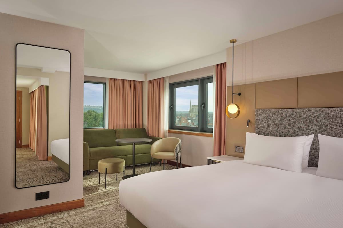 A hotel room with a large bed, a sofa, a small table, a chair, and a large mirror. There is a window with curtains and a view of a building outside.