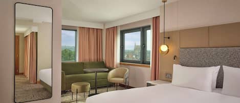 Habitación de hotel con una cama grande, un sofá, una mesita, una silla y un espejo amplio. Se aprecia una ventana con cortinas y vistas a un edificio.