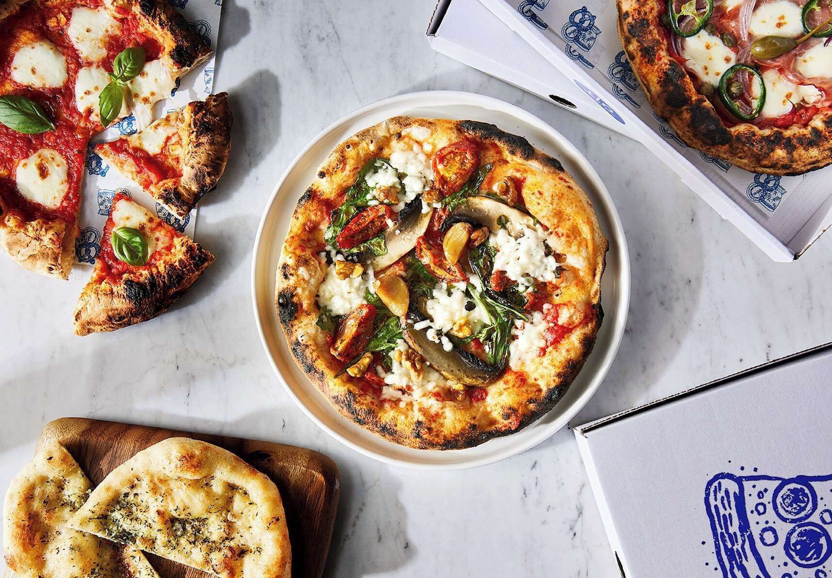 Pizza met diverse toppings, een stuk pizza met een basilicumblaadje, en een stuk knoflookbrood op een marmeren oppervlak.