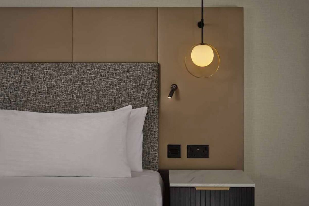 A hotel room with a bed, a headboard, a bedside table, a wall-mounted light, and electrical outlets.