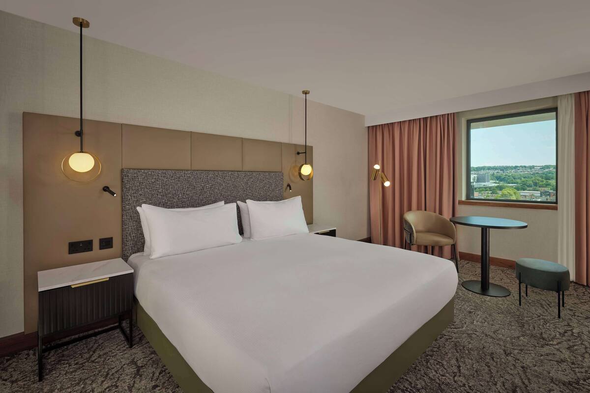 A hotel room with a large bed, a nightstand, a chair, a small table, and a view of the city through a large window.