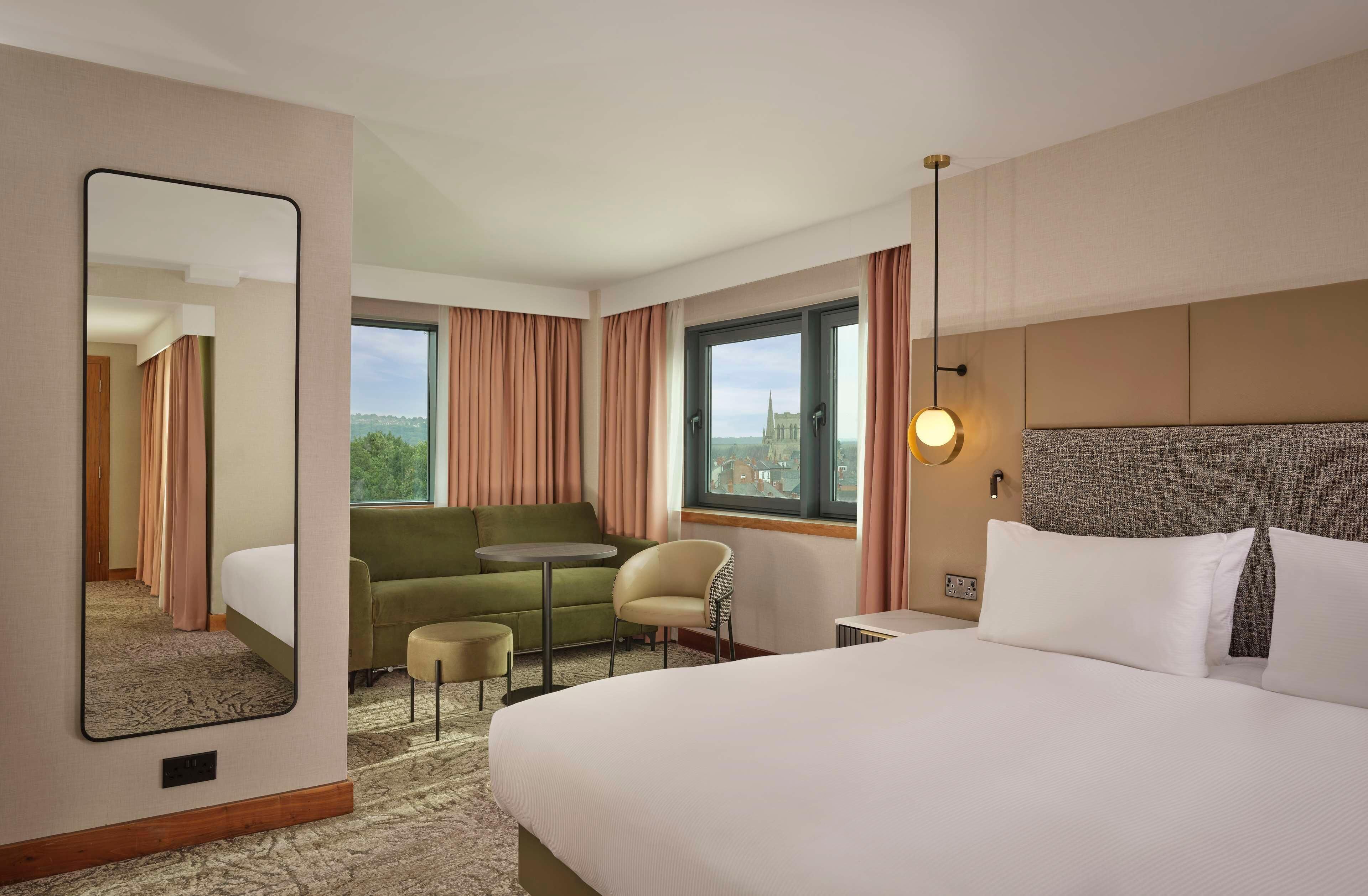 A hotel room with a bed, a sofa, a small table, a chair, a mirror, and a window with curtains.