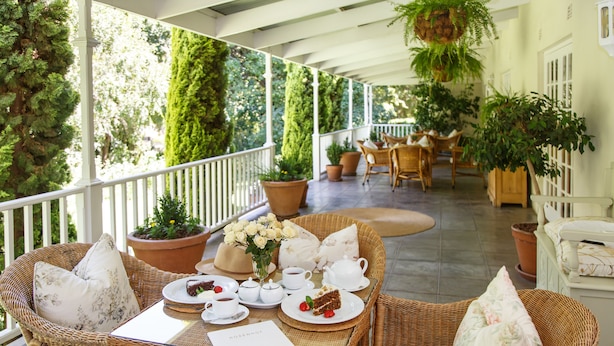 A covered outdoor dining area with wicker furniture, potted plants, and a table set for a meal. - Rosenhof Boutique Hotel (Oudtshoorn)
