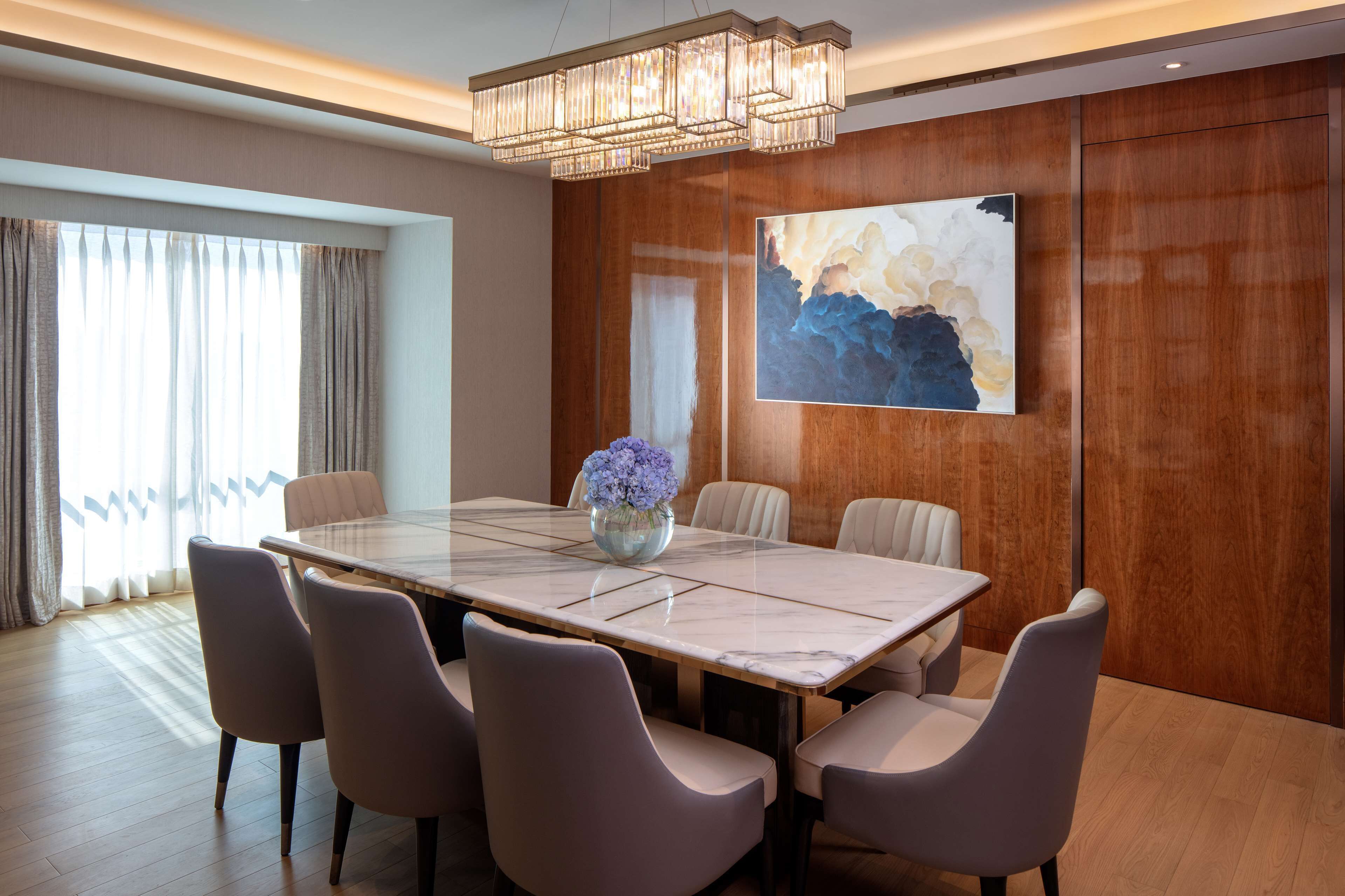 A modern dining room with a marble table, six chairs, a chandelier, and a large abstract painting on the wall.