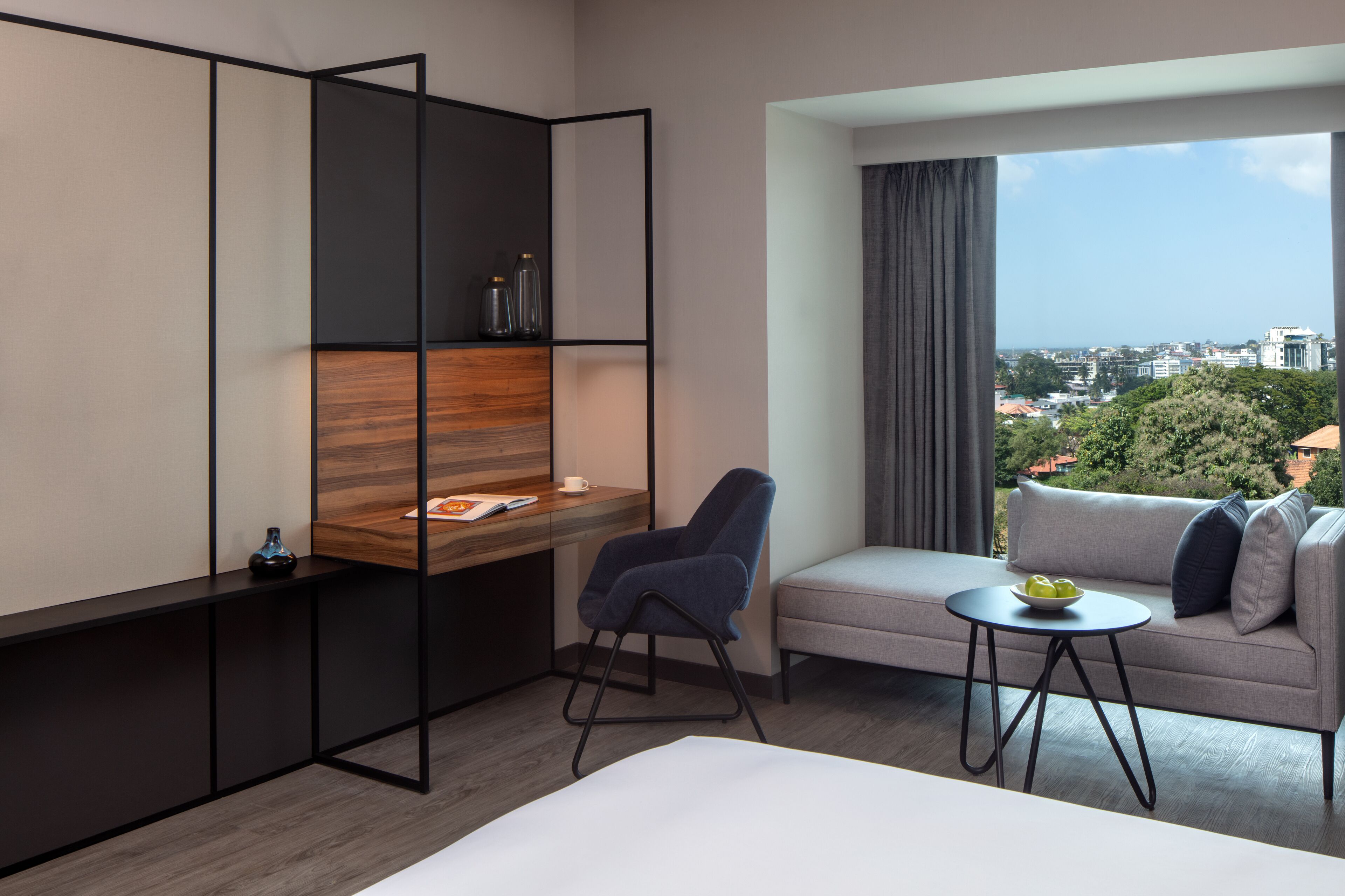 A modern hotel room with a bed, a desk with a chair, a small table with a bowl, and a sofa.