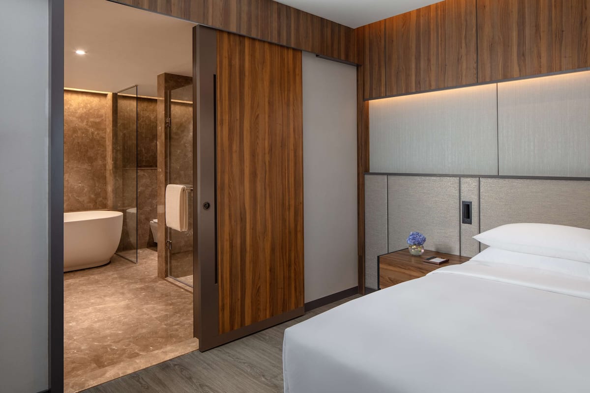 Une chambre d’hôtel moderne avec un grand lit, une armoire en bois, une salle de bain avec baignoire et une table de chevet agrémentée d’un vase.