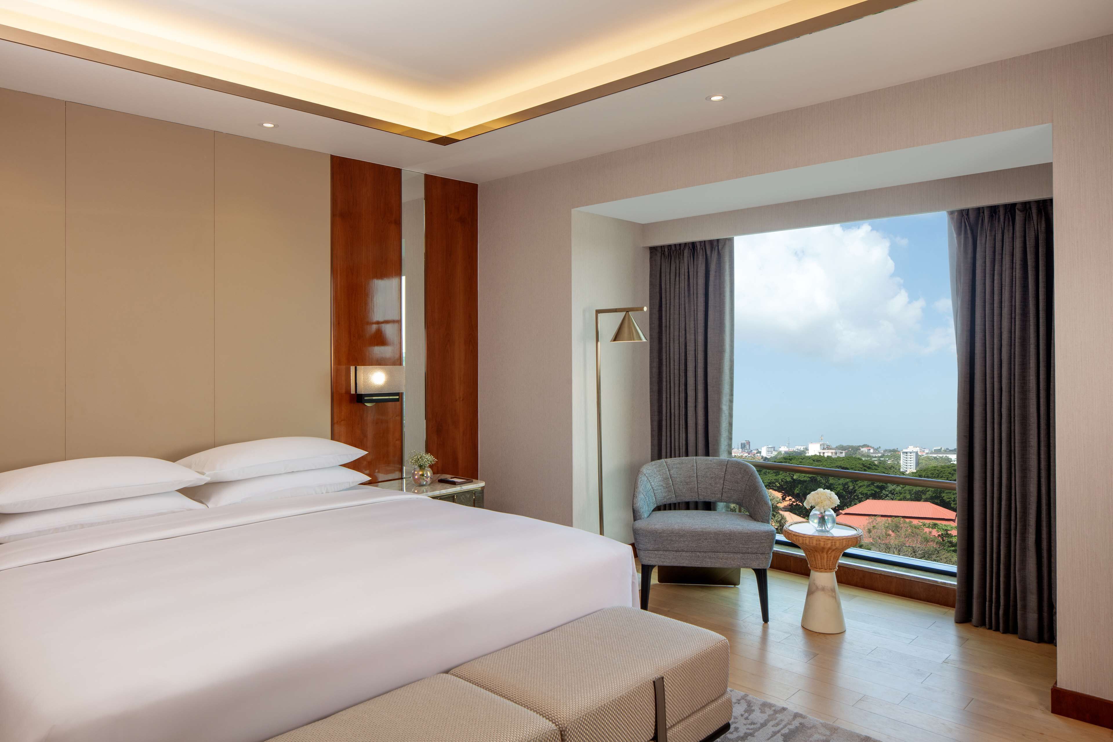A modern hotel room with a large bed, a seating area, a side table, and a view of the city through large windows.