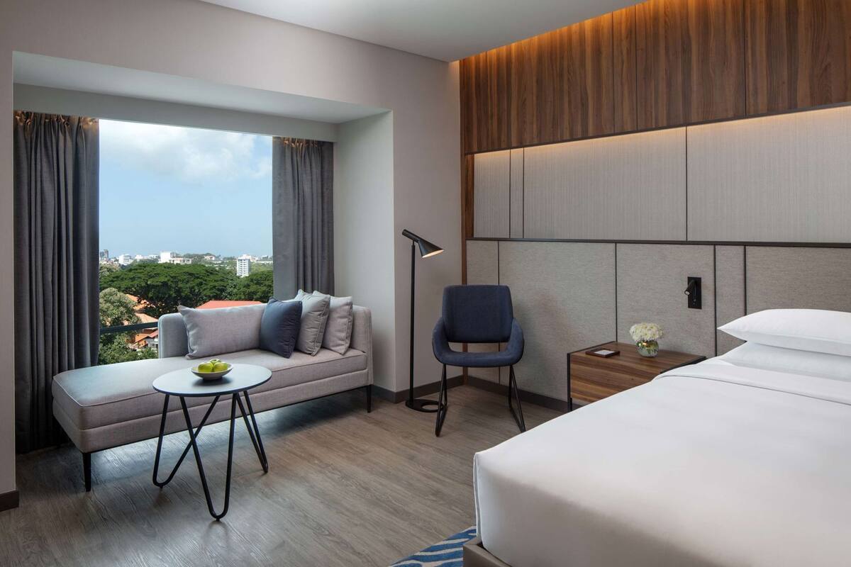 Une chambre d’hôtel avec un canapé, un fauteuil, une petite table, une table de chevet avec une lampe, un lit et une vue sur la ville.