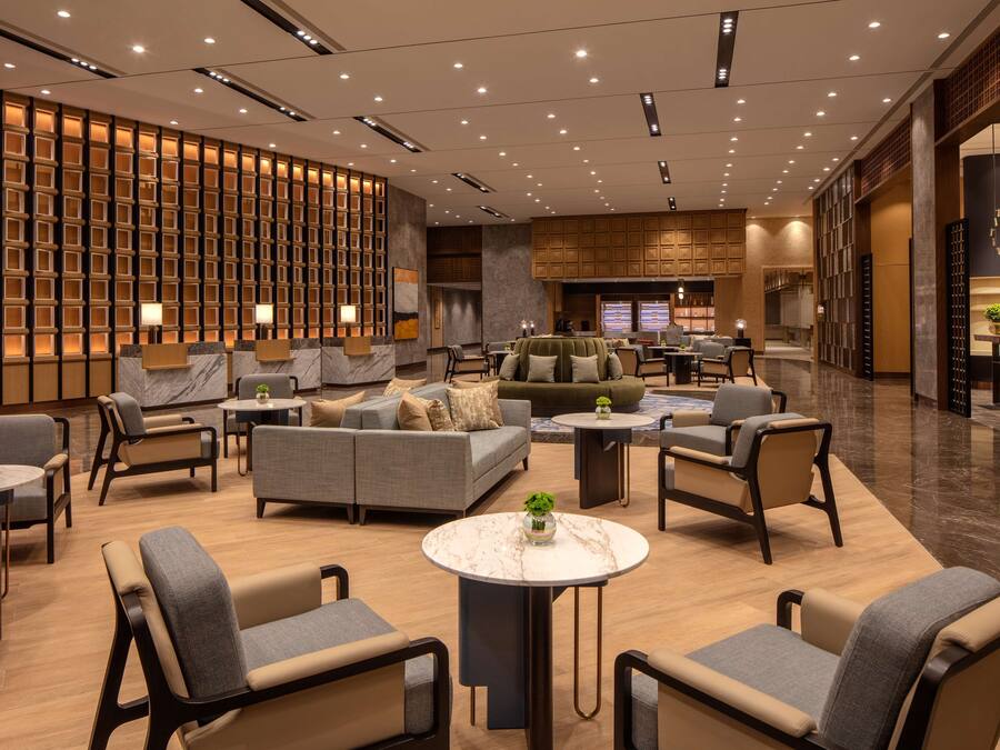 A modern hotel lobby with a seating area, a reception desk, and a kitchen visible in the background.
