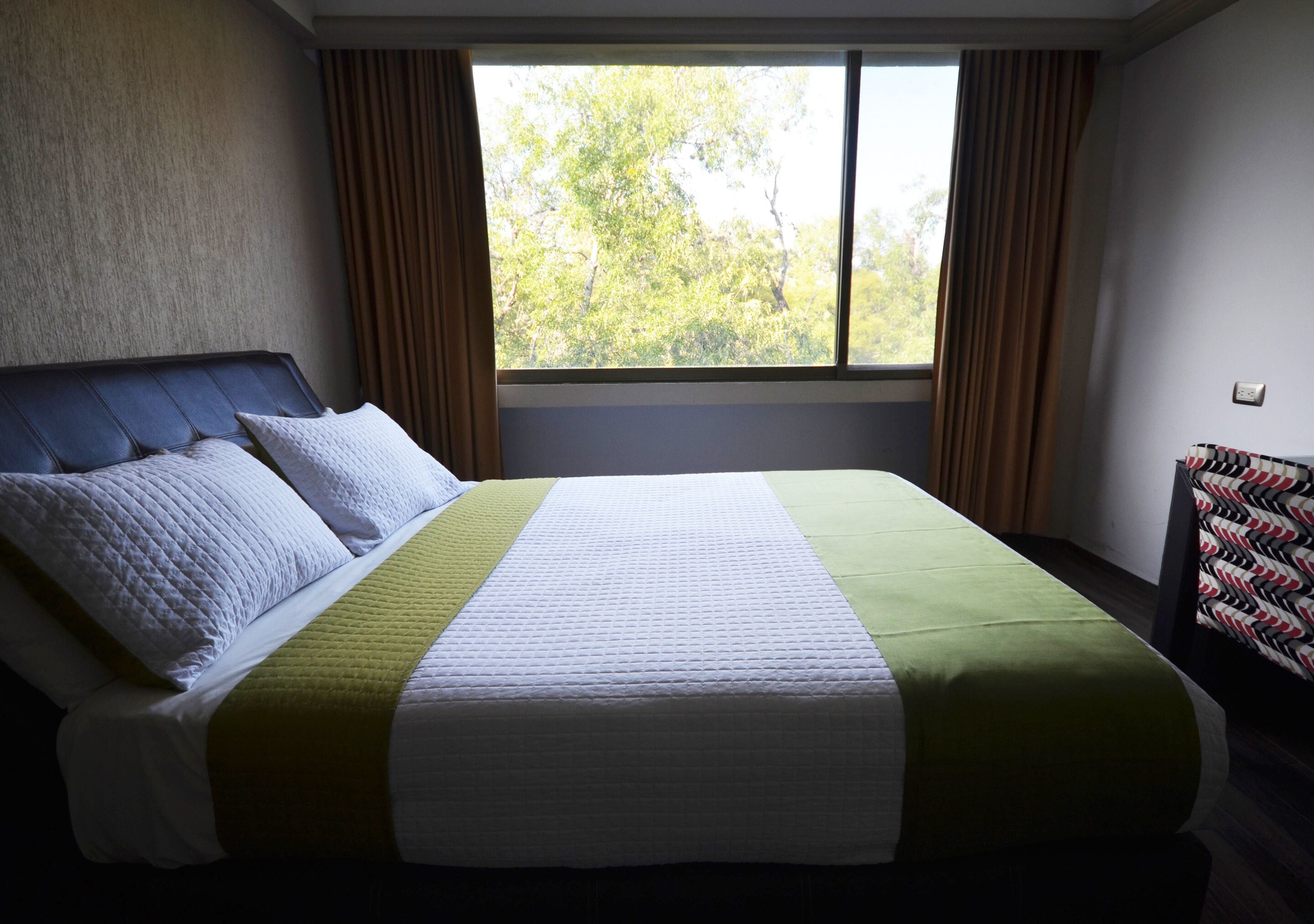 A neatly made bed with a headboard and pillows, a window with curtains, and a wall socket.