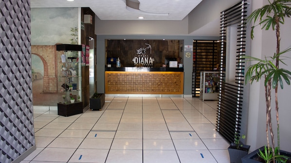 Un área de recepción con un mostrador de madera, un cartel en la pared que dice "DIANA HOTEL DEL BOSQUE" y un piso de baldosas con flechas direccionales.