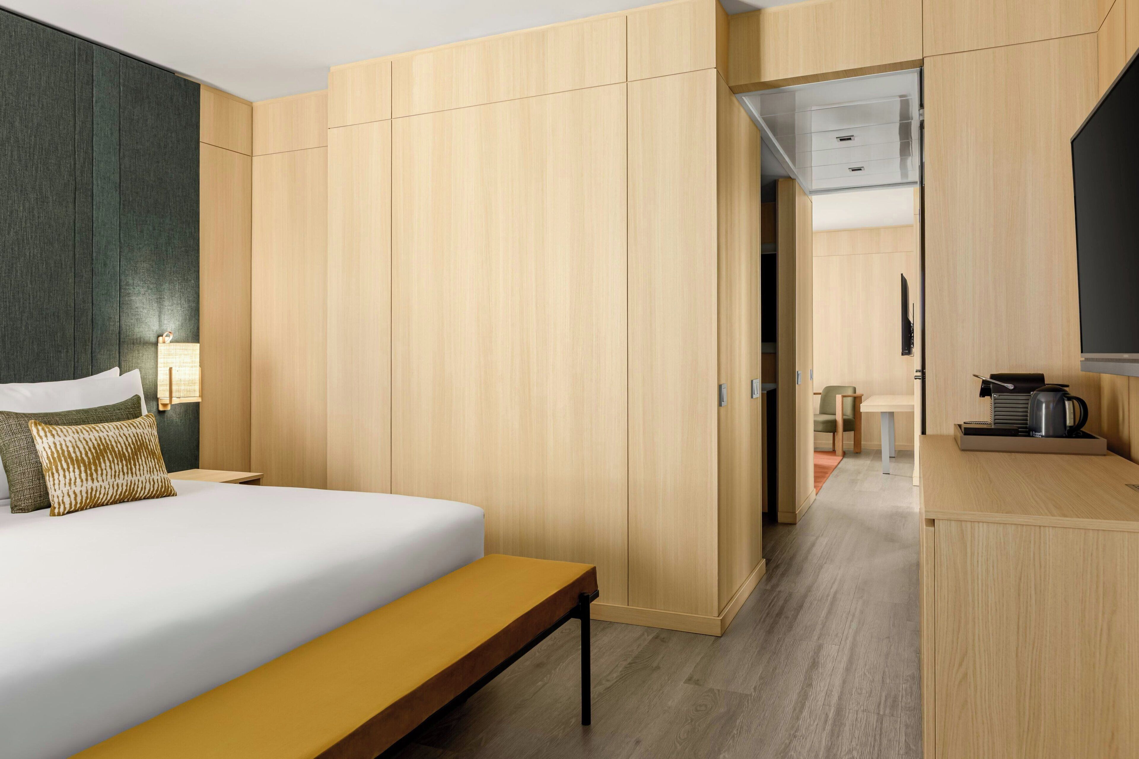 A hotel room with a bed, a bench, wooden wardrobe, a television, and a kitchenette area.