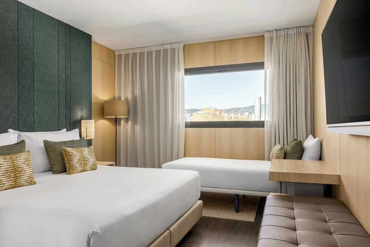 A hotel room with a bed, two bedside tables, a sofa, a window with curtains, and a view of the city.