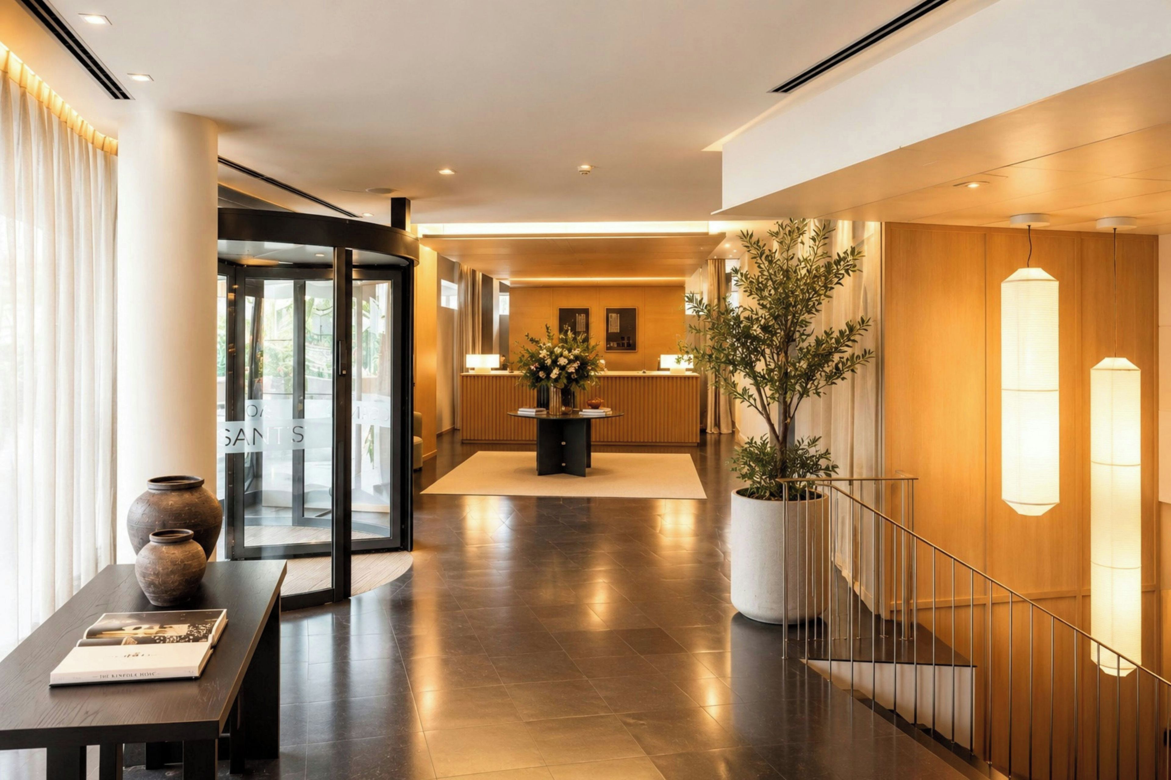Vestíbulo de hotel con mostrador de recepción, una planta en maceta, una escalera y puerta de cristal.