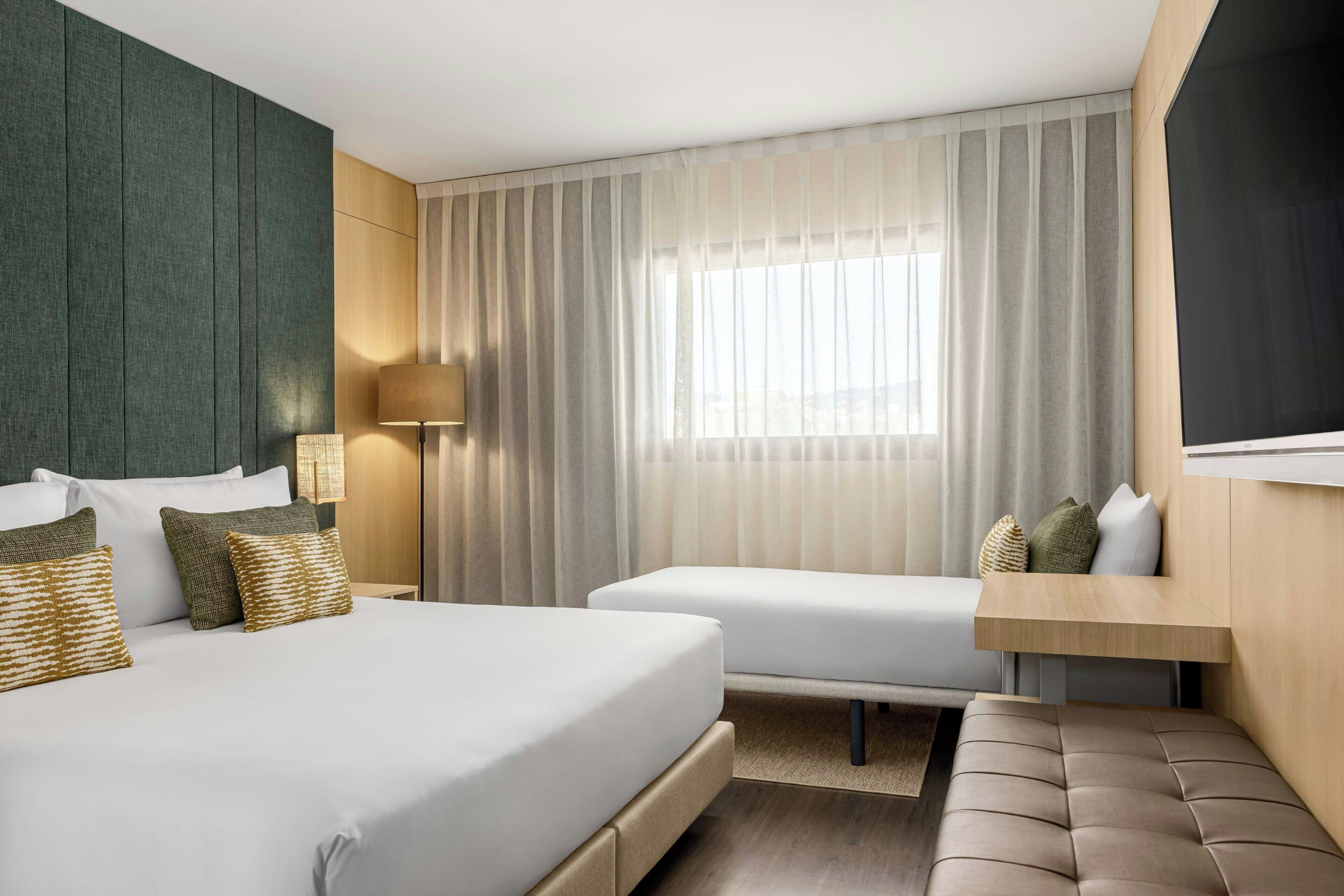 A hotel room with a bed, a sofa, a small table, a lamp, and a large window with curtains.