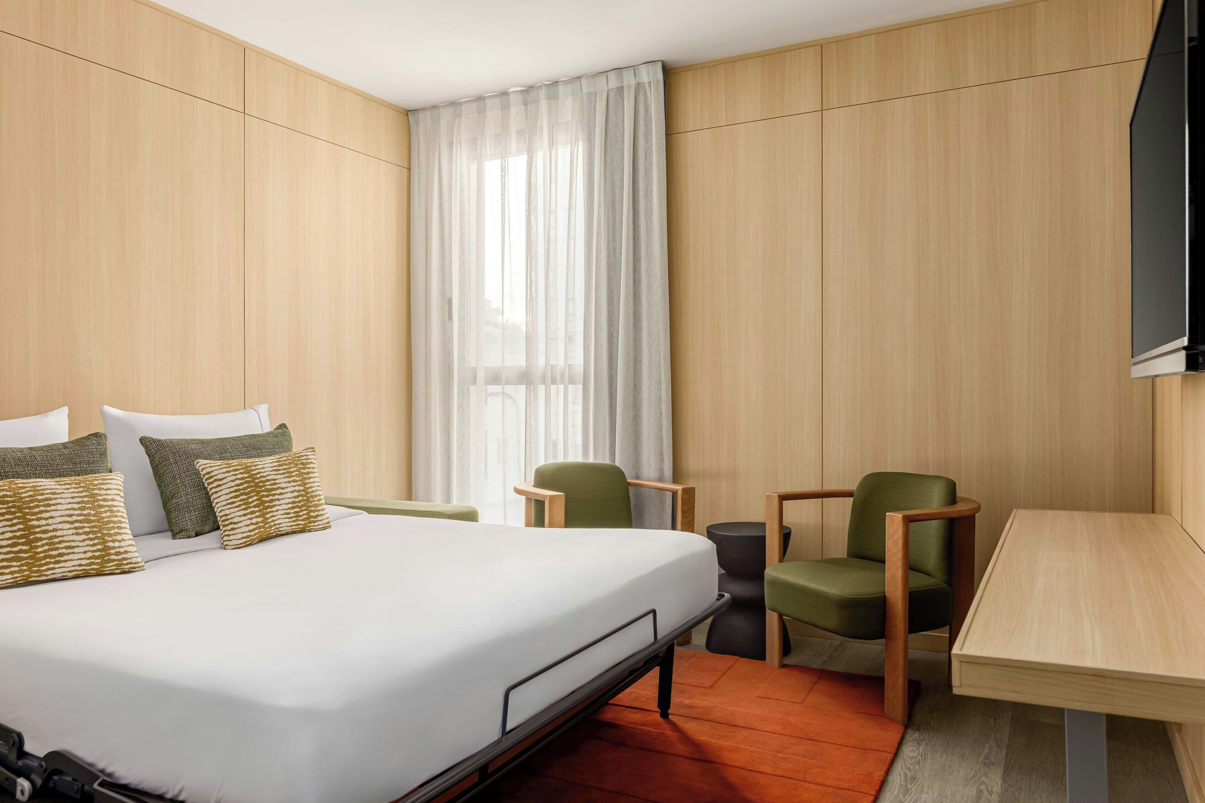 A hotel room with a bed, two chairs, a small table, and a TV mounted on the wall.