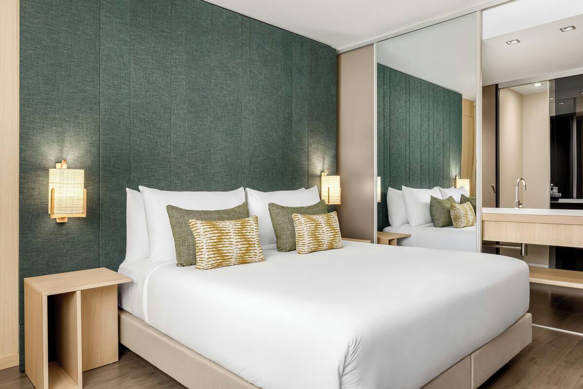 A modern hotel room with a large bed, bedside tables, a mirror, and a bathroom visible through an open door.
