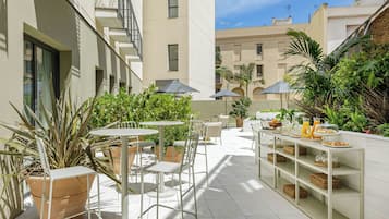 An outdoor patio area with tables and chairs, surrounded by potted plants and a buffet table with food and drinks.