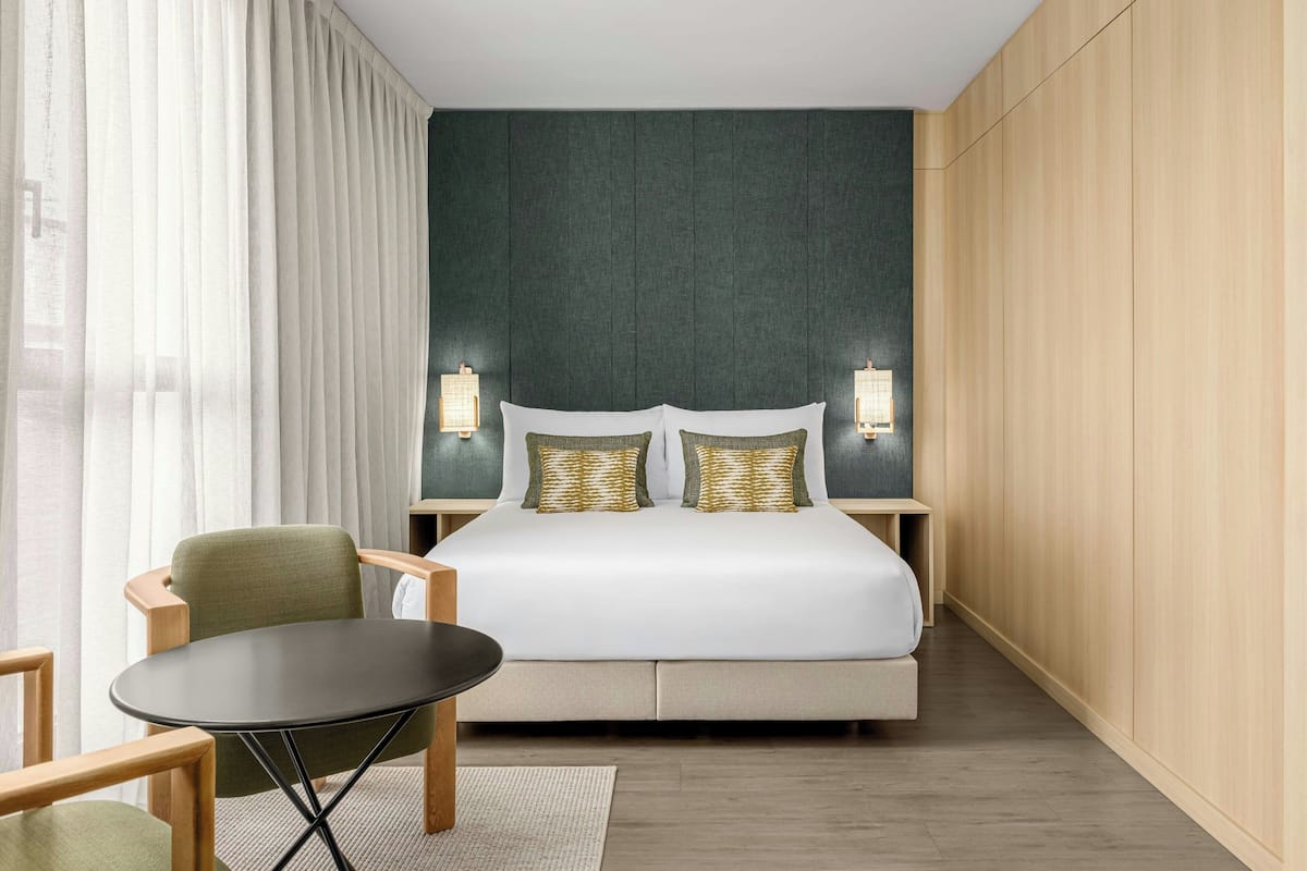 A hotel room with a bed, two nightstands, a chair, a table, and a view of the city through large windows.