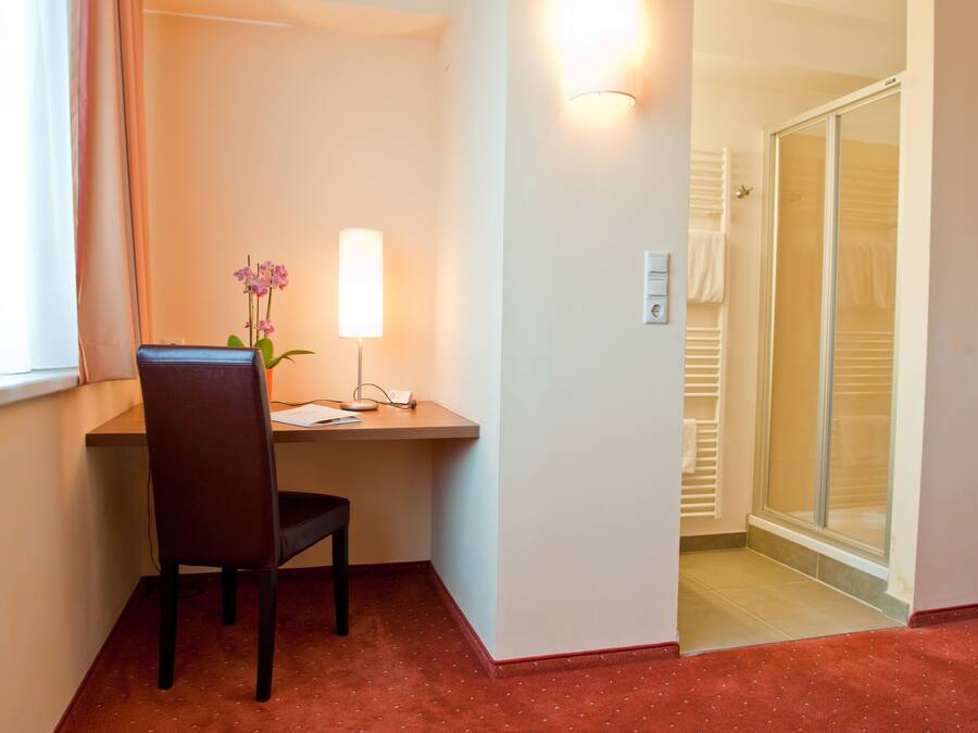 Habitación de hotel con escritorio, silla y una pequeña mesa con lámpara y un jarrón de flores. Cuenta con un área de ducha con puerta de vidrio y un perchero para toallas.